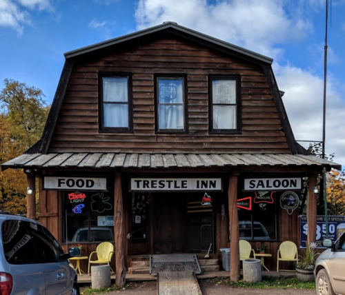 A rustic wooden building with a sign reading "Trestle Inn Saloon," featuring outdoor seating and neon lights.