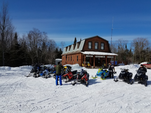 A snowy landscape with several snowmobiles parked in front of a rustic wooden lodge under a clear blue sky.