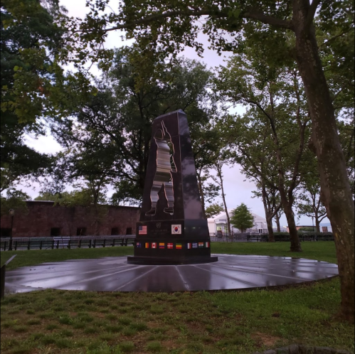 A tall, dark monument with flags at its base, surrounded by trees in a park setting.