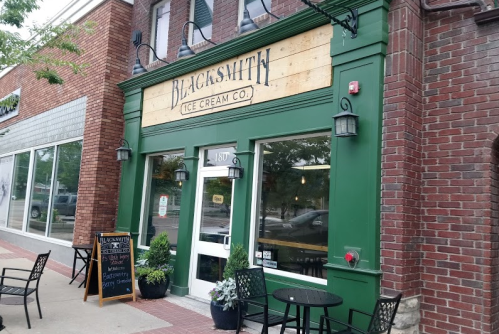Exterior of Blacksmith Ice Cream Co., featuring a green facade, wooden sign, and outdoor seating.