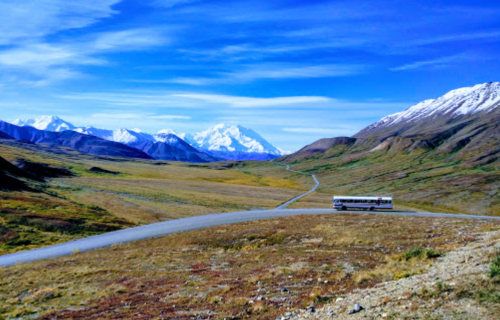 A bus travels along a winding road through a vast, mountainous landscape under a clear blue sky.
