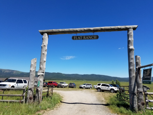 Entrance to Flat Ranch with a wooden archway, surrounded by parked cars and a clear blue sky. Mountains in the background.