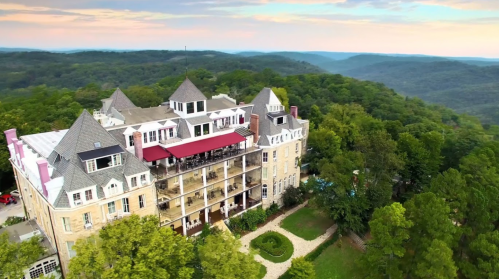 Aerial view of a large, elegant mansion surrounded by lush green trees and rolling hills under a colorful sky.