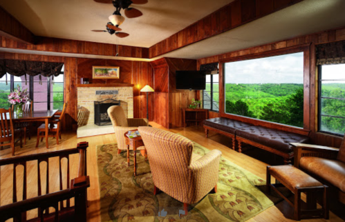 Cozy living room with wooden walls, a fireplace, and large windows showcasing a green landscape outside.
