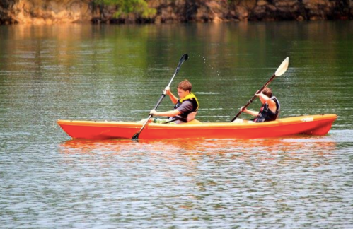 Two people paddle an orange kayak on calm water surrounded by greenery.