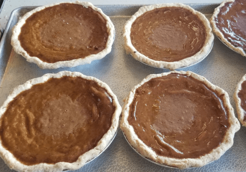 Six unbaked pies with brown filling in flaky crusts, arranged on a baking tray.