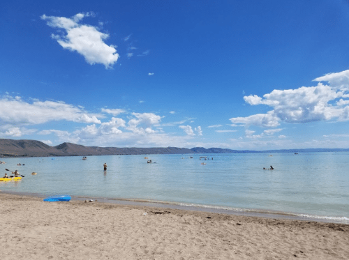 A sunny beach scene with people swimming and floating in calm water, surrounded by mountains and a blue sky with clouds.