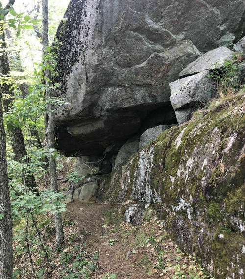 A rocky outcrop with large boulders overhanging a narrow trail, surrounded by trees and greenery.