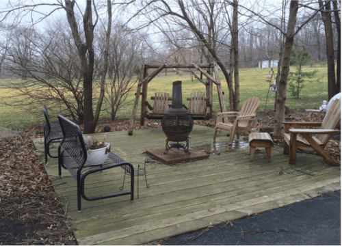 A wooden deck with seating, a chiminea, and a swing set, surrounded by bare trees and a grassy field.