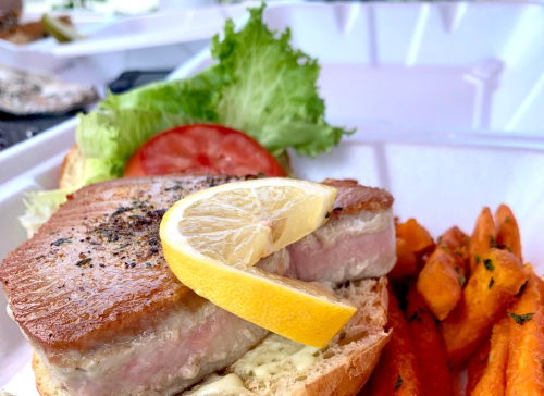 A sandwich with seared tuna, lettuce, tomato, and a lemon slice, served with sweet potato fries on the side.