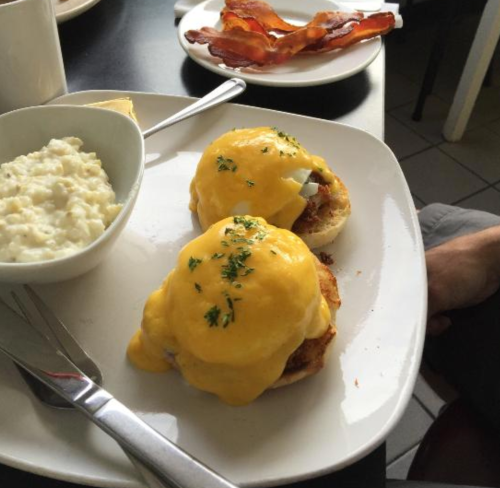 A plate with two eggs Benedict topped with hollandaise sauce, a side of cottage cheese, and crispy bacon.