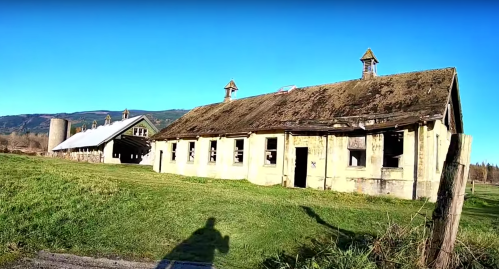 Abandoned buildings with weathered roofs and green grass under a clear blue sky, surrounded by mountains.