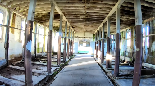 Abandoned interior of a building with exposed beams, columns, and a pathway leading to a bright exit.