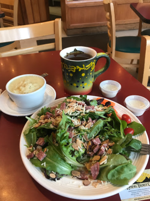 A colorful salad with greens, tomatoes, and almonds, accompanied by a cup of soup and a mug of tea on a red table.