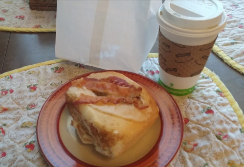 A plate with a glazed pastry topped with bacon, next to a coffee cup on a patterned tablecloth.