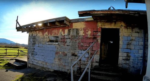 Abandoned building with peeling paint, a sloped roof, and a ramp leading to a dark entrance, set in a grassy area.