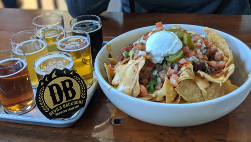 A bowl of nachos topped with cheese, tomatoes, jalapeños, and a dollop of sour cream, alongside a flight of craft beers.