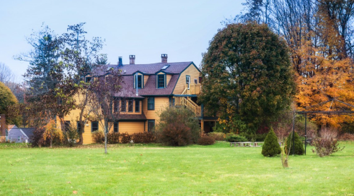 A large yellow house surrounded by trees and a grassy yard, with autumn foliage in the background.