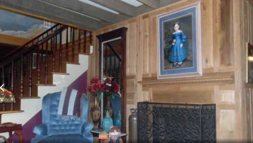Cozy living room with wooden beams, a blue chair, a fireplace, and a portrait of a girl in a blue dress on the wall.