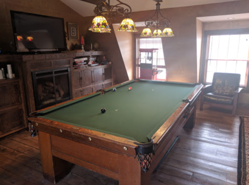 A cozy game room featuring a green pool table, stained glass lamps, and a fireplace with a TV above.
