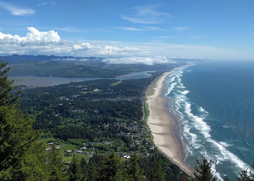 Aerial view of a coastal landscape with a sandy beach, ocean waves, and lush green hills under a blue sky.