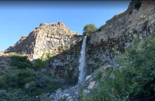 A waterfall cascades down a rocky cliff surrounded by greenery under a clear blue sky.
