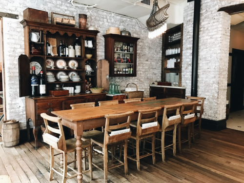 A rustic dining area featuring a wooden table, vintage chairs, and a brick wall with shelves of dishes and decor.