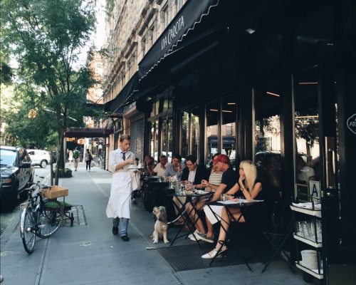 A waiter serves diners at an outdoor café, with a dog nearby and bicycles parked on the sidewalk.