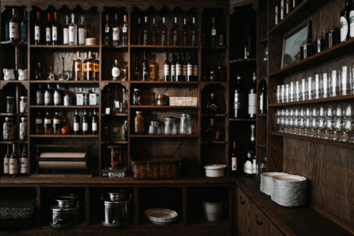 A wooden shelf filled with various bottles, glasses, and dishware in a cozy, rustic setting.