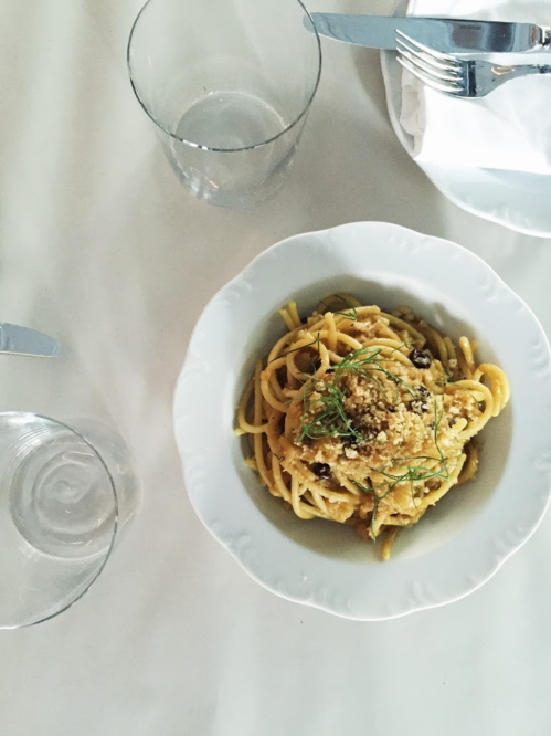 A bowl of pasta topped with breadcrumbs and herbs, surrounded by glasses and cutlery on a white table.