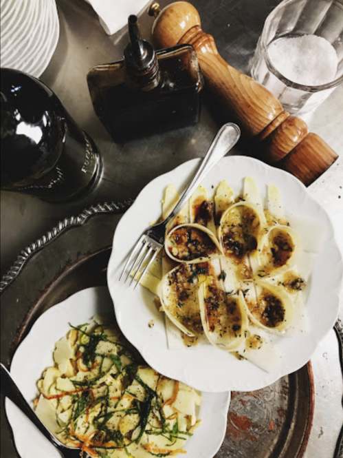 A plate of pasta with herbs and a side of sliced potatoes, accompanied by a bottle of wine and a pepper grinder.