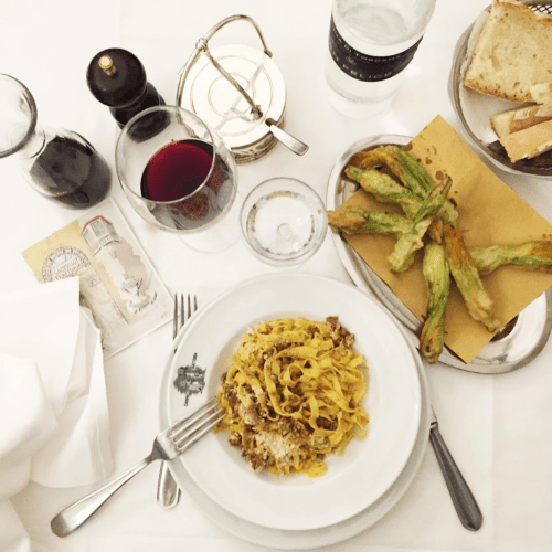 A table setting featuring pasta, stuffed zucchini flowers, red wine, water, and bread on a white tablecloth.