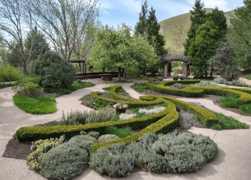 A landscaped garden with winding paths, lush greenery, and flowering plants, set against a backdrop of trees and hills.