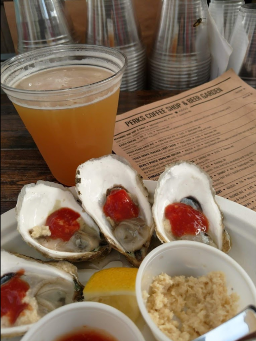 A plate of oysters topped with sauce, a lemon wedge, and a side of cocktail sauce, with a beer and menu in the background.