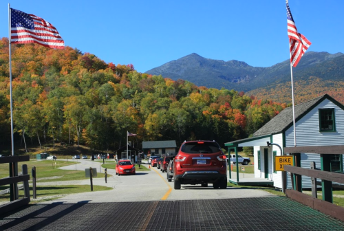 Cars lined up at a park entrance, surrounded by colorful autumn foliage and mountains under a clear blue sky.