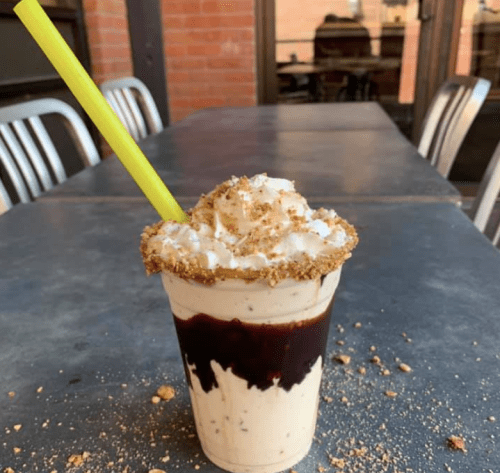 A creamy dessert drink topped with whipped cream and crushed cookies, served in a clear cup with a bright yellow straw.