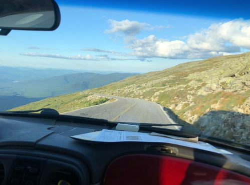 View from inside a vehicle on a winding mountain road, with lush green hills and a blue sky in the background.
