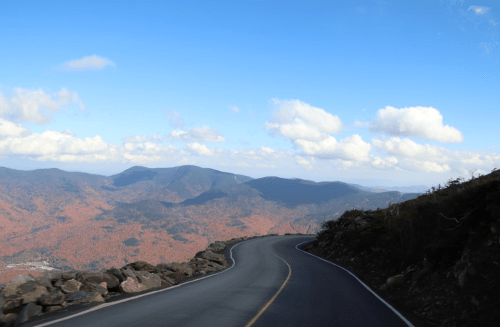 Winding road through mountains with vibrant autumn foliage under a blue sky with fluffy clouds.