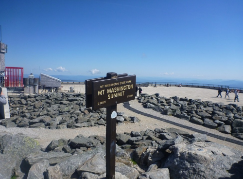 Sign at the summit of Mount Washington, with rocky terrain and visitors in the background under a clear blue sky.
