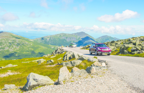 Two cars drive along a winding road surrounded by rocky terrain and green mountains under a blue sky with clouds.