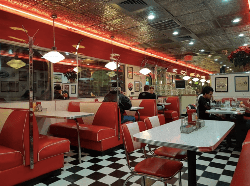 A retro diner interior with red booths, checkered floor, and warm lighting, featuring a few patrons enjoying their meals.