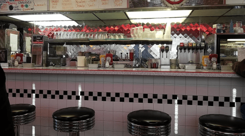 A retro diner interior featuring a counter with stools, condiments, and a mirrored backsplash with red lighting.