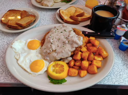 A plate of fried chicken topped with gravy, two sunny-side-up eggs, seasoned potatoes, and a lemon slice.