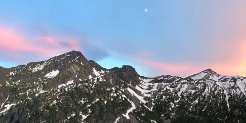 A scenic mountain landscape at dusk, featuring snow-capped peaks and a pastel sky with a crescent moon.