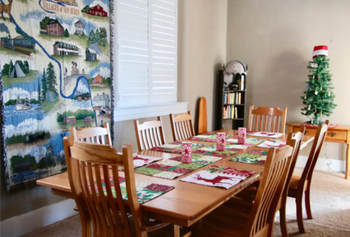 A festive dining room with a large wooden table set for Christmas, decorated with placemats and a small tree in the corner.