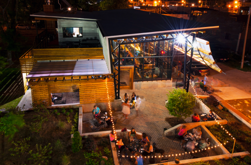 A modern restaurant with outdoor seating, surrounded by greenery, illuminated at night with warm lights.