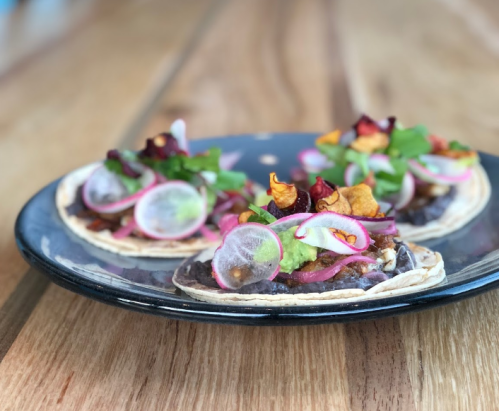 Three colorful tacos on a black plate, topped with guacamole, radishes, and crispy vegetables, on a wooden table.
