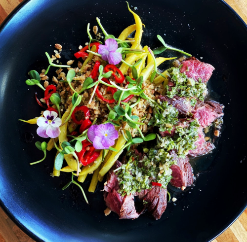 A black plate featuring sliced steak with green sauce, yellow pasta, herbs, red peppers, and edible flowers.