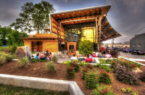 A modern building with large windows, surrounded by greenery, where people are seated outside enjoying the space.