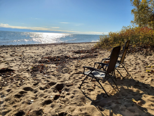 Two empty chairs on a sandy beach, overlooking a calm lake under a clear blue sky.